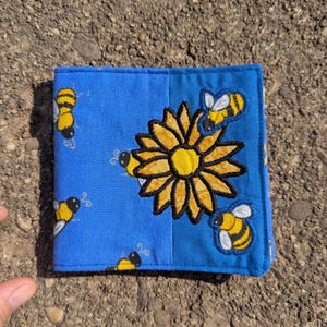 May include: A square fabric wallet in bright blue featuring a repeating pattern of small yellow and black bees. A large, central yellow sunflower with black embroidered outlines is surrounded by two additional embroidered bees on the right side.