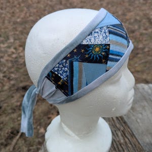 May include: A blue patchwork headband with various shades of blue fabric, including floral, striped, and celestial patterns. The headband is displayed on a white mannequin head, showcasing its design and fit. The headband has a soft, velvet-like trim and ties.