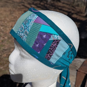 May include: A patchwork headband in shades of teal, turquoise, and purple. The headband features a variety of patterned fabrics, including paisley and geometric designs. The edges are trimmed with a solid teal fabric, and it has long ties.