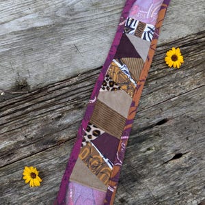 May include: A close-up of a fabric strap with a patchwork design. The strap is made of different fabrics, including corduroy, leopard print, and paisley. The strap is a deep burgundy color with a pink border.
