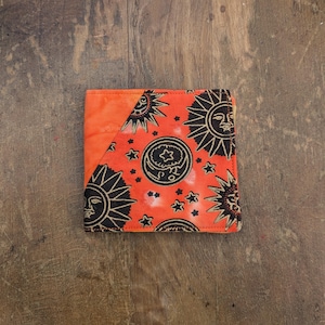 May include: A small orange wallet with a black and gold sun and moon pattern. The wallet is made of fabric and has a flap closure.