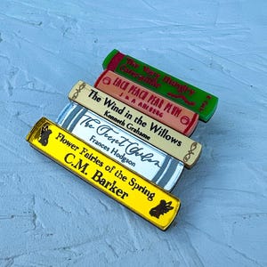 May include: A stack of five miniature books with colourful covers. The titles are "The Very Merry Mishaps of Catermelon", "Each Peach Pear Plum", "The Wind in the Willows", "The Secret Garden", and "Flower Fairies of the Spring".