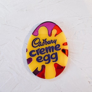 May include: A yellow, red and purple plastic egg-shaped ornament with the words "Cadbury Creme Egg" in purple lettering.