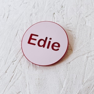 May include: A round, light pink object with the name "Edie" cut out in a dark red font. The object has a dark red border and is set against a textured white background.