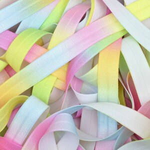 May include: A close-up of a pile of colorful elastic ribbon. The ribbon is ombre and features shades of pink, blue, yellow, and white.