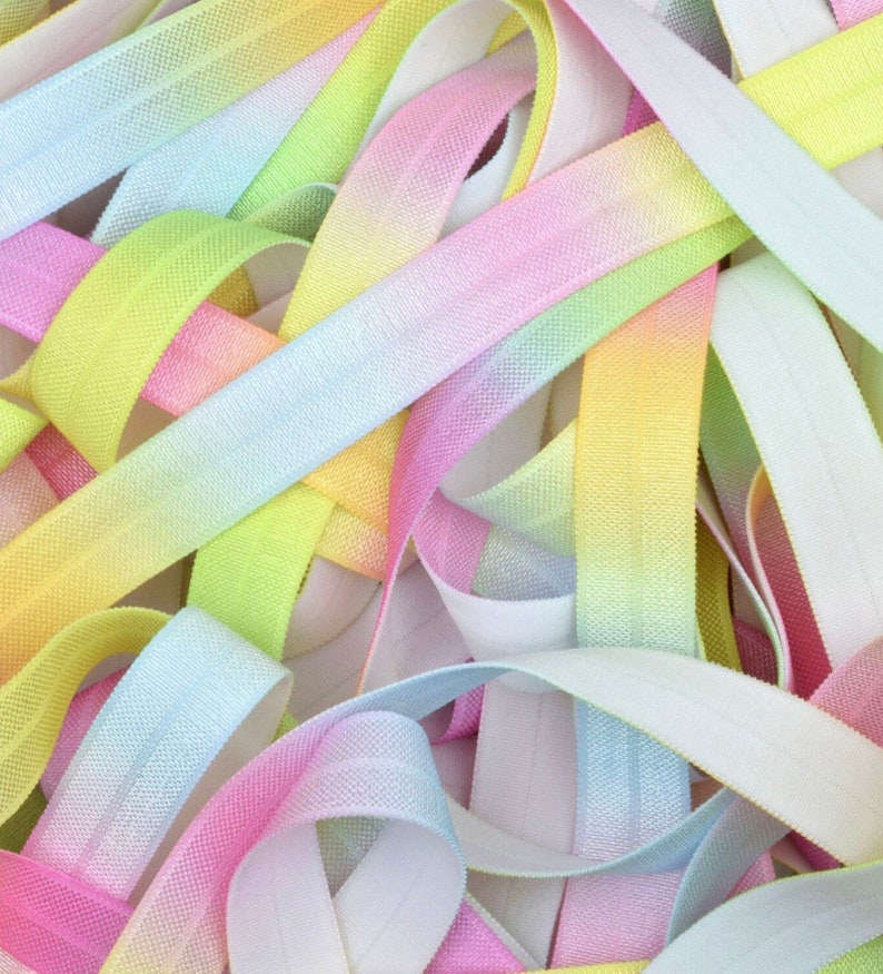 May include: A close-up of a pile of colorful elastic ribbon. The ribbon is ombre and features shades of pink, blue, yellow, and white.