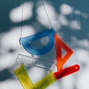 以下が含まれることがあります： 透明なプラスチック製の定規と分度器でできたネックレス。ネックレスは青い分度器、透明な定規、黄色の分度器、オレンジ色の定規でできています。ネックレスは銀色のチェーンからぶら下がっています。