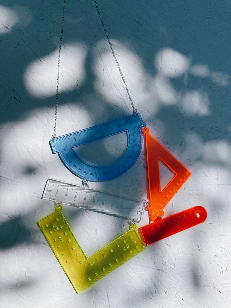 以下が含まれることがあります： 透明なプラスチック製の定規と分度器でできたネックレス。ネックレスは青い分度器、透明な定規、黄色の分度器、オレンジ色の定規でできています。ネックレスは銀色のチェーンからぶら下がっています。