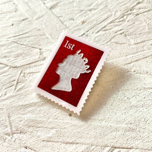 May include: A white framed red postage stamp with a silver silhouette of a woman's profile wearing a crown. The stamp says "1st" in the top left corner.