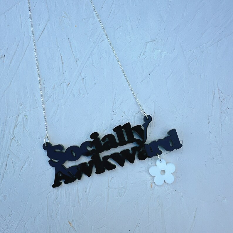 May include: A black necklace with a silver chain and a pendant that says "Socially Awkward" with a white flower charm.