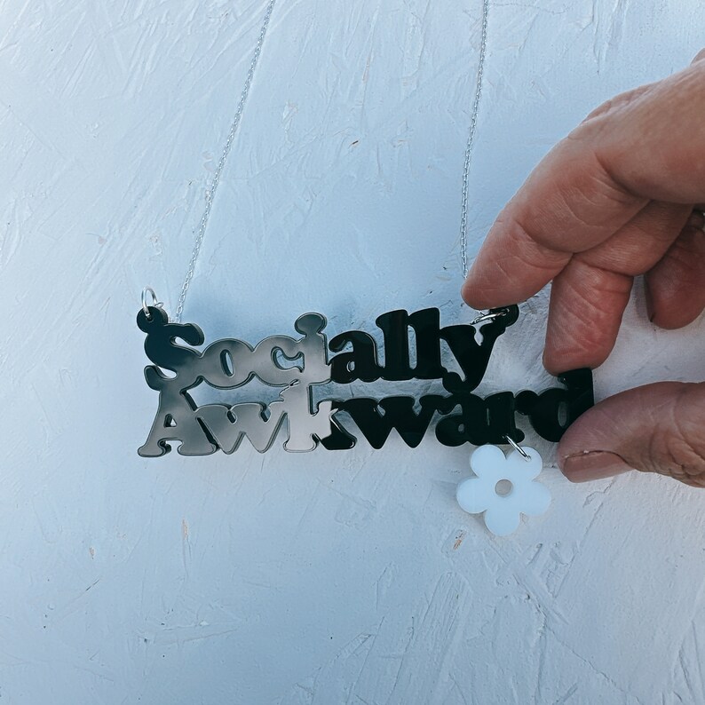May include: A silver chain necklace with a black and white acrylic pendant that reads "Socially Awkward". The pendant has a white flower charm attached to it.