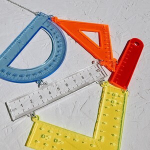 以下が含まれることがあります： 分度器、三角形、定規など、カラフルな幾何学定規のセット。定規は銀色のチェーンでつながっています。