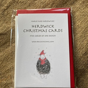 May include: A pack of five Christmas cards featuring a Herdwick sheep wearing a red hat and scarf in a snowy scene. The cards are packaged in a red envelope and are available from www.beccafielding.com.