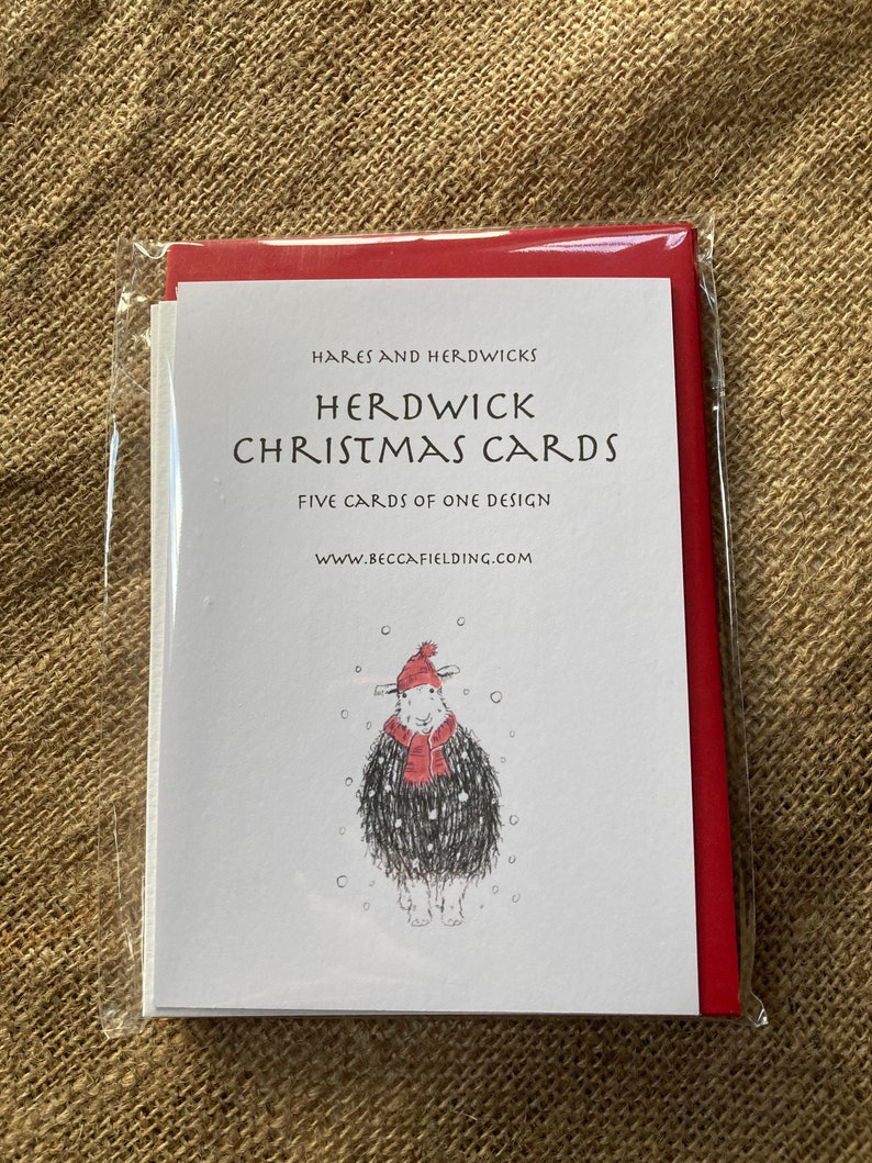 May include: A pack of five Christmas cards featuring a Herdwick sheep wearing a red hat and scarf in a snowy scene. The cards are packaged in a red envelope and are available from www.beccafielding.com.