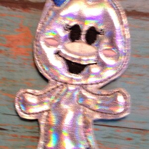 May include: A whimsical, silver-colored applique in the shape of a cartoon character. It features a blue bow, black eyes, and a smiling mouth. The character's arms and legs are outlined in silver with blue accents on the feet.