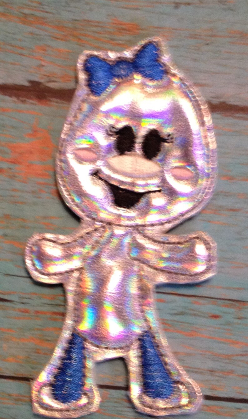 May include: A whimsical, silver-colored applique in the shape of a cartoon character. It features a blue bow, black eyes, and a smiling mouth. The character's arms and legs are outlined in silver with blue accents on the feet.