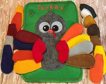 May include: A green felt board featuring a gray turkey with large eyes and a red wattle. The word Turkey is embroidered in orange at the top. The set includes numerous detachable felt feathers in various colors including red, orange, yellow, blue, brown, gray, and white, designed to be attached to the turkey body.
