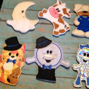 May include: A collection of colorful, embroidered felt appliques featuring whimsical characters. Includes a smiling moon, a cow, a dog in overalls, a cat with a violin, a snowman, and a sparkly figure with a bow.