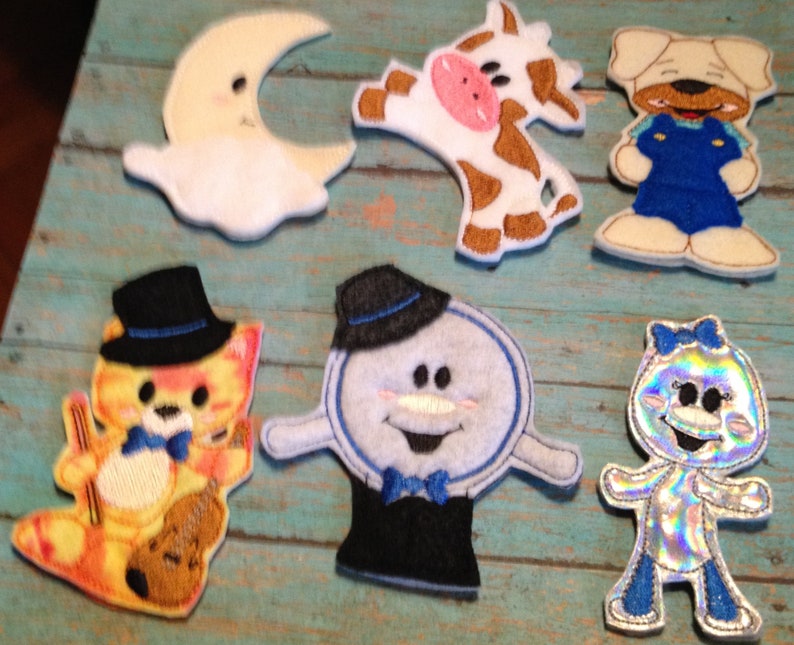 May include: A collection of colorful, embroidered felt appliques featuring whimsical characters. Includes a smiling moon, a cow, a dog in overalls, a cat with a violin, a snowman, and a sparkly figure with a bow.