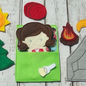 May include: A felt camping scene with a green tent, a sun, a tree, a campfire, and a crescent moon. A girl is in a green sleeping bag. There are also three red felt circles and a flashlight.