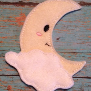 May include: A felt moon with a smiling face and a cloud. The moon is a light yellow color with a black eye, a pink blush, and a stitched mouth. The cloud is white and stitched to the moon.