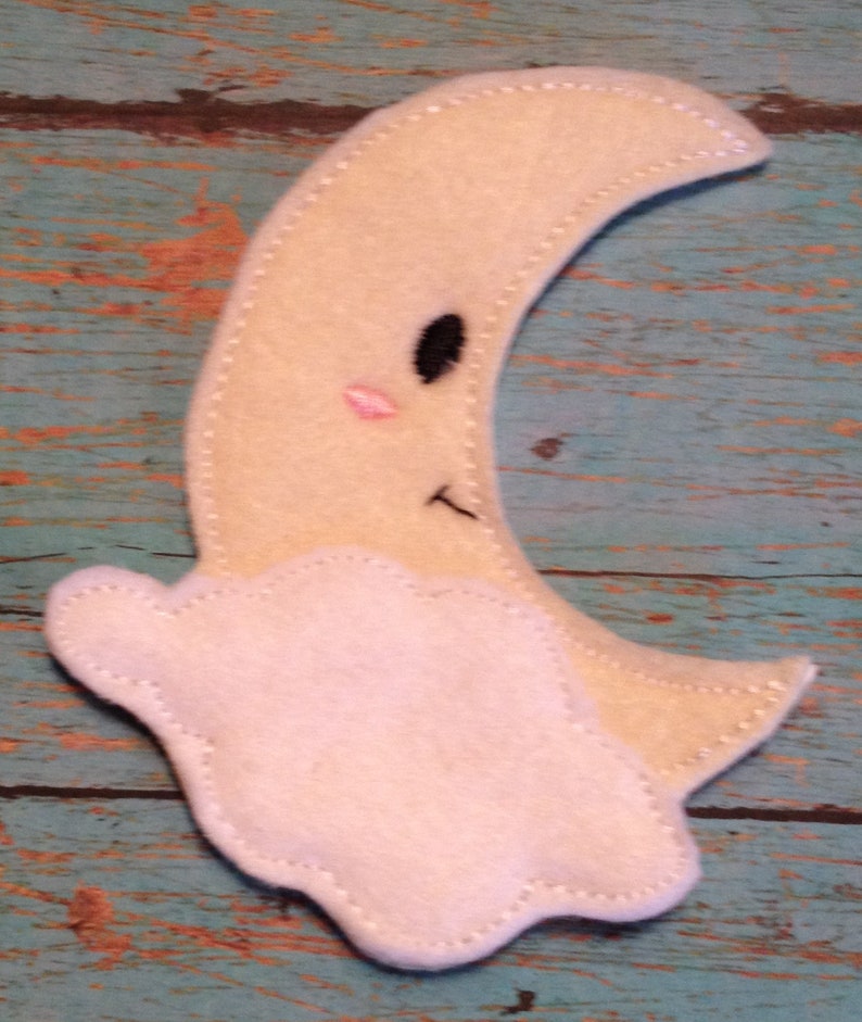 May include: A felt moon with a smiling face and a cloud. The moon is a light yellow color with a black eye, a pink blush, and a stitched mouth. The cloud is white and stitched to the moon.