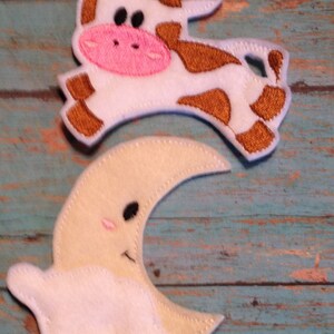 May include: Felt cow and moon-and-cloud shapes. The cow is white with brown spots and a pink nose. The moon is cream-colored with a smiling face and a white cloud. The shapes are likely for a nursery or children's craft project.