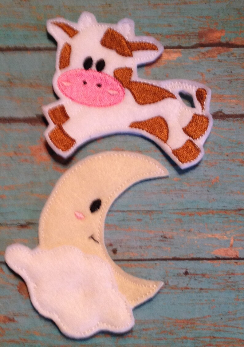 May include: Felt cow and moon-and-cloud shapes. The cow is white with brown spots and a pink nose. The moon is cream-colored with a smiling face and a white cloud. The shapes are likely for a nursery or children's craft project.