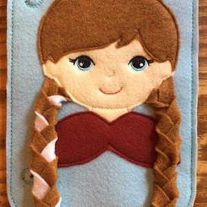 May include: A felt doll face with brown hair in braids, blue eyes, and a red top, stitched onto a light blue felt background. The doll has a smiling expression and is designed with a button detail.