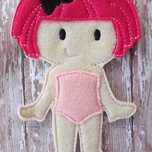 May include: A felt doll with a pink leotard, pink hair, and a black bow. The doll has a smiling face with black eyes and a stitched mouth. The doll's arms and legs are outstretched, and the edges are stitched with white thread.
