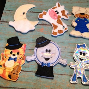 May include: A collection of colorful felt appliques featuring various cartoon characters. Includes a smiling moon, a cow, a dog in overalls, a cat playing a violin, a snowman, and a holographic figure with a blue bow.