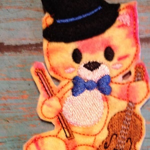 May include: Embroidered patch of a cat playing a violin. The cat is wearing a black top hat and a blue bow tie. The cat is yellow, orange, and pink with black eyes and a black nose. The violin is brown.