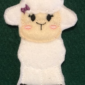 Puede incluir: Un t&iacute;tere de dedo de fieltro blanco con forma de oveja, con cara color crema, ojos bordados en negro, nariz y boca peque&ntilde;as en negro, mejillas bordadas en rosa y un peque&ntilde;o lazo morado en la cabeza. La base tiene pies bordados en negro.