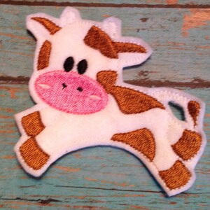 May include: An embroidered patch in the shape of a cartoon cow. The cow is white with brown spots, a pink snout, and black eyes. The patch is designed to look like the cow is running or jumping.