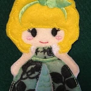 Puede incluir: Una aplicaci&oacute;n de fieltro de una mu&ntilde;eca con cabello amarillo brillante, una diadema verde claro y mejillas rosadas. La mu&ntilde;eca viste un vestido con un patr&oacute;n floral verde oscuro y negro sobre una base azul claro, con costuras visibles en los bordes.