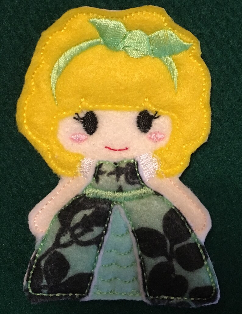 Puede incluir: Una aplicaci&oacute;n de fieltro de una mu&ntilde;eca con cabello amarillo brillante, una diadema verde claro y mejillas rosadas. La mu&ntilde;eca viste un vestido con un patr&oacute;n floral verde oscuro y negro sobre una base azul claro, con costuras visibles en los bordes.
