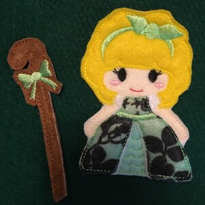 Puede incluir: Un conjunto de apliques de fieltro que incluye una mu&ntilde;eca con cabello amarillo brillante, una diadema verde y un vestido verde con estampado floral negro, acompa&ntilde;ado de un cayado de pastor de fieltro marr&oacute;n con un lazo verde y bordes cosidos.