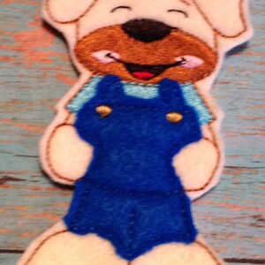 May include: A felt dog-shaped ornament with a smiling face, wearing a blue overall. The dog has a tan face and ears, with a brown nose and mouth. The overall has gold-colored buttons. The ornament is likely handmade.