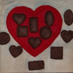 May include: A red felt heart with several dark brown felt chocolates of various shapes, including hearts, squares, and ovals. The chocolates have white stitched details. The arrangement suggests a Valentine's Day theme.