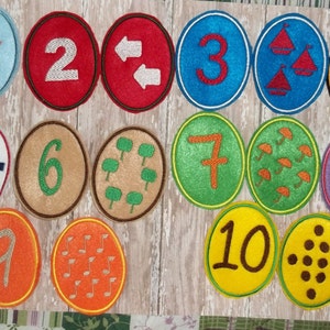 May include: A collection of colorful, embroidered patches featuring numbers 1 through 10, each with a unique design. The patches are in various colors, including blue, red, green, and orange, with white borders. The designs include stars, arrows, and hearts.