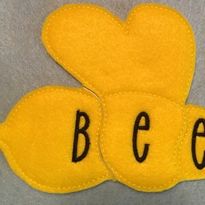 May include: A yellow felt applique shaped like a stylized bee with rounded wings and body segments. The word bee is embroidered in black lowercase letters across the three connected yellow felt sections with visible stitching.