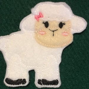 Puede incluir: Una aplicaci&oacute;n de fieltro blanco con forma de cordero que presenta una cara de color crema, ojos bordados en negro, un peque&ntilde;o lazo rosa en la cabeza, mejillas bordadas en rosa y pezu&ntilde;as negras. Los bordes tienen costuras blancas visibles.