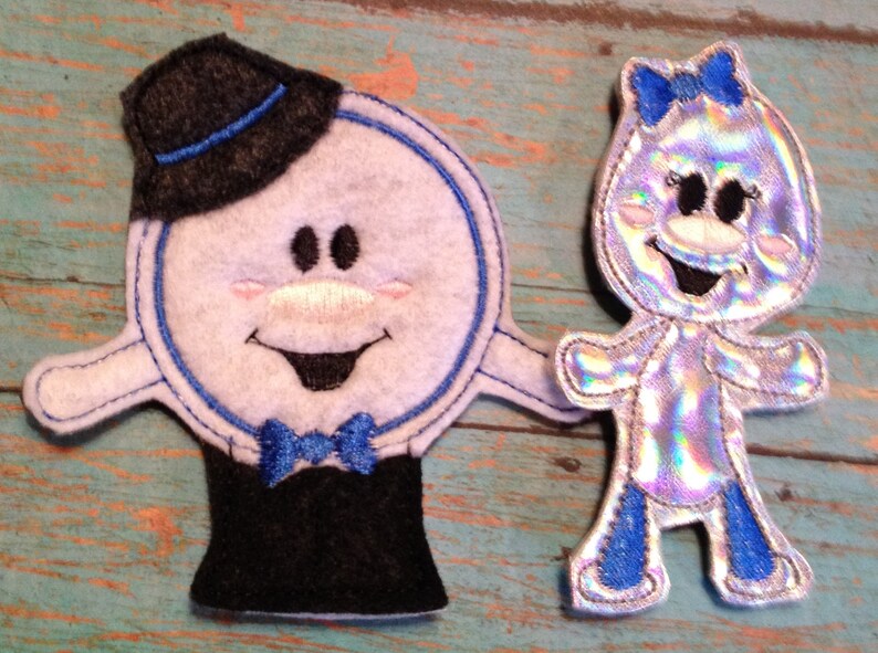 May include: Two whimsical felt figures. One is a white circle with a black top hat and bow tie, outlined in blue. The other is a silver, iridescent figure with a blue bow and accents. Both have smiling faces.