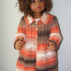 May include: A doll wearing a striped, knitted cardigan with a brown, orange and white colour scheme. The cardigan has a collar and buttons down the front.