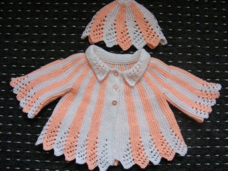 May include: A knitted baby cardigan and hat set in pink and white stripes with a scalloped edge. The cardigan has a button closure and a collar.