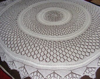 May include: A large, circular, white crocheted tablecloth. The intricate design features concentric circles with openwork patterns, including diamond shapes and a scalloped edge. The delicate, handmade textile is displayed on a brown surface.