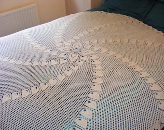 May include: A cream-coloured, crocheted bedspread with a central floral motif. The circular design radiates outwards, with openwork leaf-shaped details around the edges. The bedspread is displayed on a bed with a teal bedsheet.