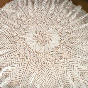 May include: A white, circular, crocheted doily with a detailed floral center and a scalloped edge. The intricate openwork design features a spiral pattern in the center, surrounded by geometric and floral motifs. Small pink accents are visible along the edge.