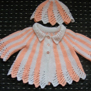 May include: A white and peach striped knitted baby cardigan with a white collar and peach trim. The cardigan has a scalloped edge and is paired with a matching peach and white knitted hat.