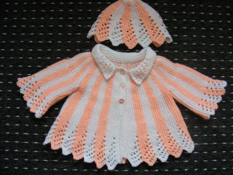May include: A white and peach striped knitted baby cardigan with a white collar and peach trim. The cardigan has a scalloped edge and is paired with a matching peach and white knitted hat.